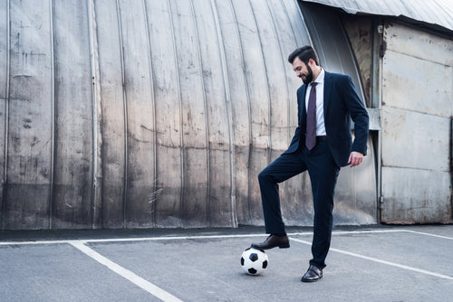 Man in a suit playing with a soccer ball in a parking lot, symbolizing a balance of professionalism and play