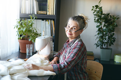 Smiling older woman with glasses and a measuring tape, sitting by a sewing machine with books and plants in the background